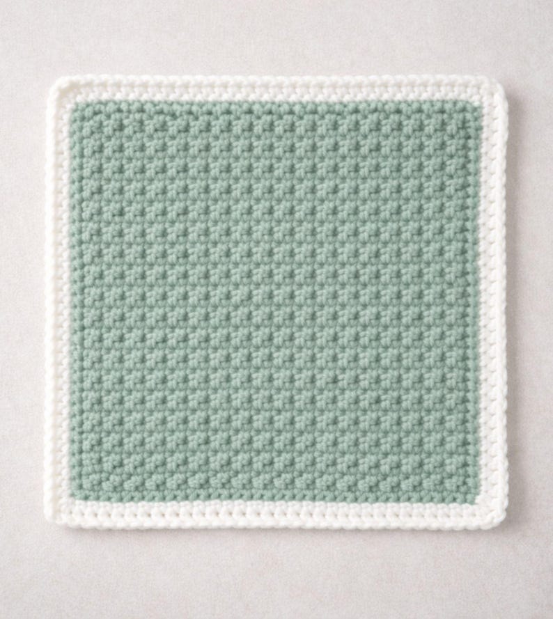 May include: A square crochet coaster featuring a textured sage green center panel with a consistent, repeating stitch pattern. The square is framed by a solid white border made of a smooth, uniform crochet stitch that outlines the entire perimeter.