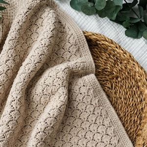 May include: A beige crochet blanket featuring a repeating shell stitch pattern. The textile has a soft, textured appearance with a consistent light tan colour throughout the fabric, showcasing the intricate loops and knots of the yarn construction.