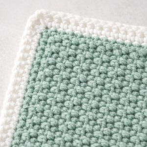 May include: A square crochet textile featuring a sage green center with a textured, bumpy stitch pattern. The piece is framed by a thick, smooth white border made of simple crochet stitches, creating a clean contrast between the two colors.