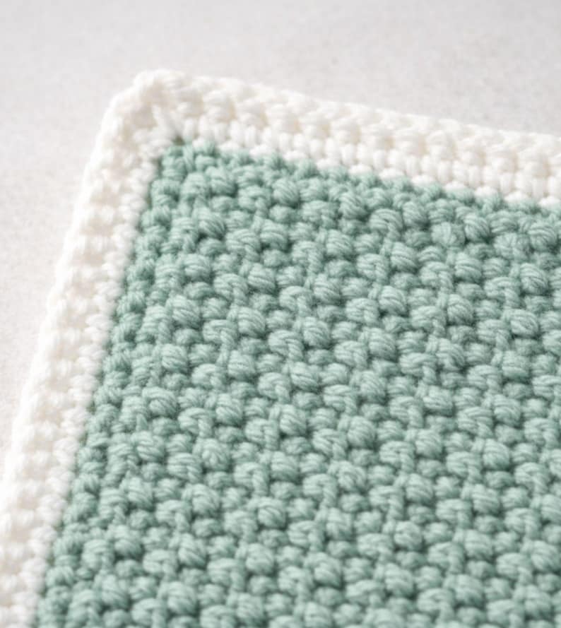 May include: A square crochet textile featuring a sage green center with a textured, bumpy stitch pattern. The piece is framed by a thick, smooth white border made of simple crochet stitches, creating a clean contrast between the two colors.