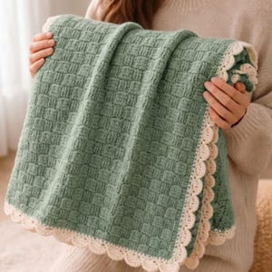 May include: A sage green crocheted blanket featuring a textured basketweave stitch pattern throughout the center. The edges of the blanket are finished with a decorative, scalloped cream-colored crochet border that frames the entire piece.