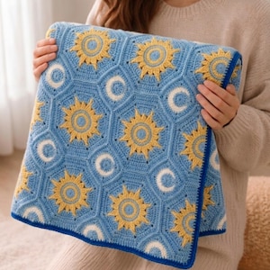 May include: A blue crocheted blanket with a celestial pattern of yellow suns and white moons. The blanket, with a blue border, is held by a person. The handmade blanket is a cozy home decor item, perfect for adding a touch of warmth.
