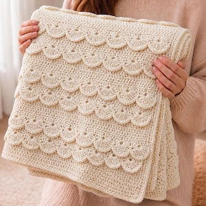 May include: A cream-coloured crocheted blanket featuring a repeating pattern of horizontal rows of textured, fan-shaped scalloped edges. The textile is crafted with a dense, uniform stitch throughout, creating a layered, dimensional appearance.