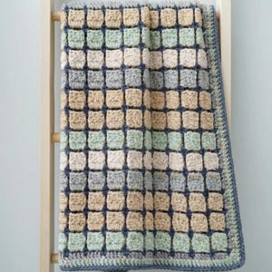 May include: A handmade crochet blanket with a textured, square pattern in shades of beige, green, gray, and blue. The blanket is draped over a wooden ladder, showcasing its detailed craftsmanship and color palette. The border is a solid blue.