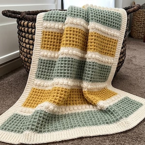 May include: A crochet blanket featuring a waffle stitch pattern in alternating blocks of sage green and mustard yellow. The blanket is bordered by cream-colored yarn and includes horizontal rows of raised bobble stitches between the colored sections.