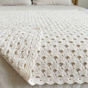 May include: A cream-colored crochet blanket featuring a repeating fan or shell stitch pattern. The textile has a textured, scalloped edge border and a consistent, intricate woven design throughout the entire surface of the rectangular piece.