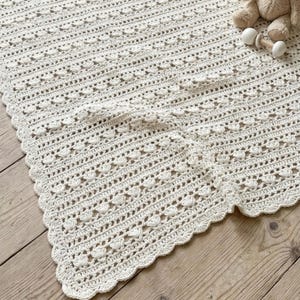 Puede incluir: Una manta de ganchillo color blanco roto con un patrón texturizado repetitivo de pequeños grupos en forma de concha y filas caladas. Los bordes tienen un acabado festoneado. Se ve un pequeño peluche color canela y un sonajero de madera blanco.