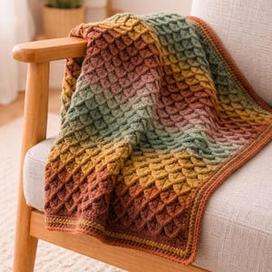 May include: A crochet blanket featuring a textured scale pattern in a gradient of colors including deep rust, mustard yellow, sage green, and soft pink. The rectangular textile has a finished border and is draped over a wooden chair.