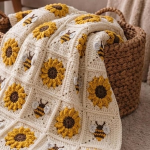 May include: A handmade crochet blanket featuring a repeating square pattern of yellow sunflowers with dark brown centers and small bees with white wings, all set against a cream-colored yarn background, draped inside a woven brown wicker basket.