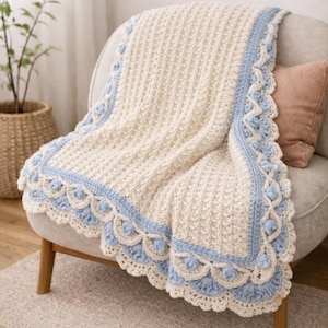May include: A cream-colored crochet blanket with a textured pattern and a light blue scalloped border. The blanket is draped over a chair, showcasing its intricate design and soft texture. A decorative pillow rests on the chair.
