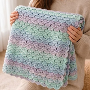 May include: A square crochet blanket featuring a repeating scalloped shell pattern in a gradient of soft pastel colours including light blue, mint green, and lavender. The textured yarn fabric is arranged in horizontal rows of overlapping semi-circles.