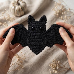 May include: A black crochet bat with two pointed ears and round black eyes. The bat is crafted with a textured yarn stitch pattern, featuring wide, outstretched wings and a small, pointed body shape, displaying a detailed handmade construction.