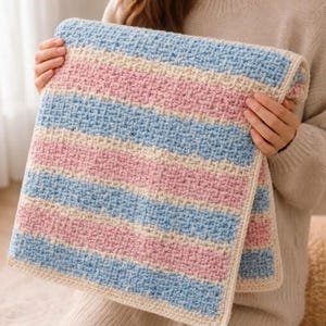 Pode incluir: Uma manta de crochê retangular com listras horizontais em cores alternadas: azul claro, rosa suave e creme. O tecido apresenta um padrão de pontos texturizado e entrelaçado, finalizado com uma borda sólida na cor creme.
