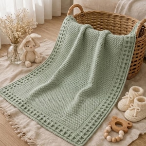 May include: A sage green baby blanket with a textured pattern and a decorative border. The blanket is folded over a wicker basket, with a crocheted bunny, baby booties, and a wooden teether nearby. The blanket is approximately 91 x 102 cm.