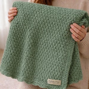May include: A sage green crocheted blanket featuring a textured stitch pattern and a scalloped edge. A small rectangular fabric label is attached to the bottom corner, displaying the text "Handmade with Love" in a clear, dark font.