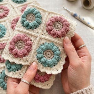 May include: Handmade crochet squares with flower designs. The squares have a cream background with pink and teal flowers. The crochet work is detailed, with a scalloped edge. The squares are held in a person's hands.