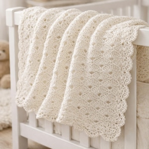 Peut inclure: Une couverture en crochet de couleur crème présentant un motif répétitif de points en éventail et un bord festonné. Le textile est drapé sur le côté d'un cadre en bois blanc, mettant en valeur le travail complexe du fil et la texture aérée.