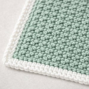 May include: A square crochet textile featuring a textured sage green center panel with a dense, raised stitch pattern. The piece is finished with a clean, white crocheted border that frames the edges, creating a distinct contrast in color.