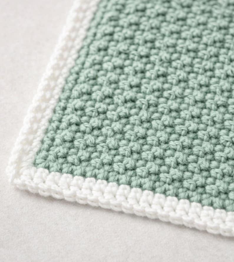 May include: A square crochet textile featuring a textured sage green center panel with a dense, raised stitch pattern. The piece is finished with a clean, white crocheted border that frames the edges, creating a distinct contrast in color.