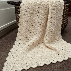 May include: A cream-colored crochet blanket featuring a repeating shell stitch pattern throughout the body and a scalloped edge border. The textile has a textured, open-work design and is draped over a dark brown woven basket.