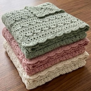 Op de afbeelding: Een stapel van drie gehaakte dekens in tinten groen, roze en crème. Elke deken heeft een geschulpte rand en een delicaat openwerkontwerp. De bovenste deken is gevouwen met een kleine, decoratieve flap.