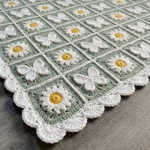 May include: A crochet blanket featuring a repeating grid pattern of white daisies with yellow centers and white butterflies on a sage green background. The blanket is finished with a scalloped white crochet border along the outer edge.