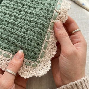 May include: A sage green crocheted textile featuring a textured stitch pattern and a scalloped cream-colored border. The edge is finished with a woven cream crochet trim threaded with a thin, smooth sage green ribbon that weaves through the border.