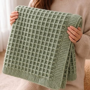 Può includere: Una coperta lavorata a maglia verde salvia con una trama a nido d'ape. La coperta è piegata, mostrando il motivo a punto dettagliato e il colore morbido e tenue. I bordi hanno un bordo a coste.