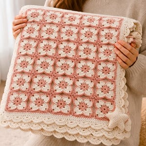 Puede incluir: Una manta cuadrada de ganchillo con un diseño floral en tonos rosa y crema. La manta tiene un borde festoneado y un adorno en forma de estrella. La manta hecha a mano está siendo sostenida por una persona.