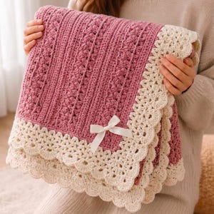 May include: A rectangular crochet blanket featuring a dusty rose pink centre with vertical textured stitch patterns and a wide, cream-coloured scalloped border. A small, simple white ribbon bow is attached to the front of the pink section.