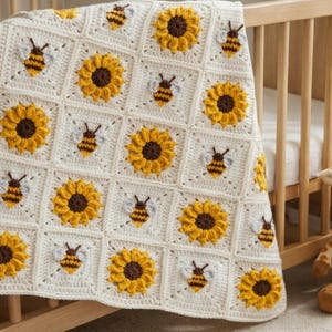 May include: A cream-colored crochet blanket featuring a repeating square pattern. Each square displays either a textured yellow sunflower with a dark brown center or a small bee with yellow and brown stripes and light gray wings.