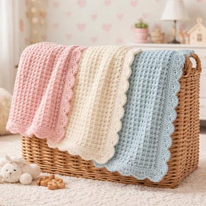 May include: Three crocheted baby blankets in pink, cream, and blue with scalloped edges are displayed in a wicker basket. The blankets have a textured stitch pattern. A white stuffed bunny and wooden toys are in the foreground.