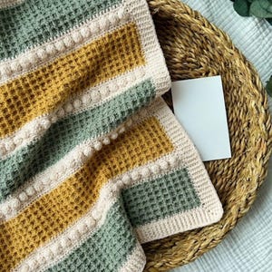 May include: A crocheted blanket featuring a waffle stitch pattern with alternating horizontal stripes in sage green and mustard yellow. The design includes rows of raised bobble stitches in cream, bordered by a solid cream-coloured crochet edge.