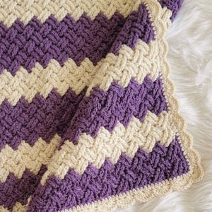 May include: A hand-crocheted blanket featuring a textured basketweave stitch pattern in alternating diagonal stripes of deep purple and cream yarn. The edges of the blanket are finished with a decorative scalloped border in a matching cream color.