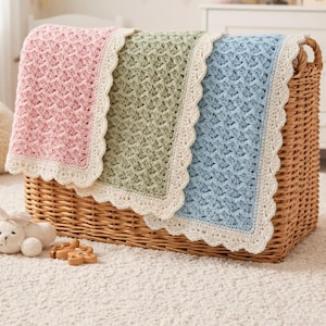 May include: Three crocheted baby blankets in pink, green, and blue, each with a white scalloped edge, are displayed in a wicker basket. The blankets have a textured pattern. A white stuffed animal and wooden toys are in the foreground.