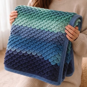 May include: A square crochet blanket featuring a textured scale pattern in an ombre gradient of colors. The design transitions from light mint green at the top to medium blue, and finally to a deep navy blue at the bottom, finished with a blue border.