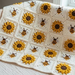 May include: A crochet blanket composed of square motifs featuring alternating patterns of yellow and brown sunflowers and small bees with white wings, all set against a cream-colored background, creating a repeating grid design throughout.
