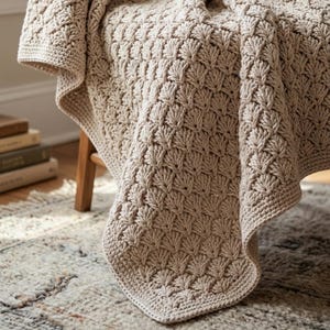May include: A beige, hand-crocheted blanket with a textured, fan-like pattern. The blanket is draped over a wooden chair, showcasing its intricate design and soft texture. The neutral color and detailed stitching suggest a cozy, handmade item.