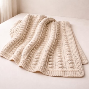 Può includere: Una coperta all'uncinetto strutturata color crema con un motivo ripetuto di colonne verticali e disegni quadrati. La coperta è piegata, mostrando la sua dettagliata lavorazione artigianale e la sua morbida consistenza. La coperta è realizzata in un materiale morbido e caldo.