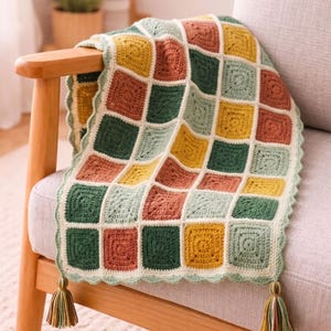 Op de afbeelding: Een handgemaakte gehaakte deken met een patchwork-ontwerp, gedrapeerd over een stoel. De deken heeft vierkanten in groen, geel, oranje en crème. Het heeft een geschulpte rand en kwastjes op de hoeken. Een decoratief item voor in huis.