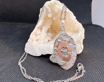 May include: A handcrafted silver pendant necklace with a textured, organic design. The pendant features a copper-colored oval accent and is suspended from a delicate silver chain. The necklace is displayed against a white, crystalline background.