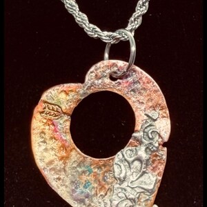 May include: A heart-shaped pendant with a silver chain. The pendant has a textured, multi-colored surface with copper, silver, and hints of blue. A leaf design is visible on the pendant.