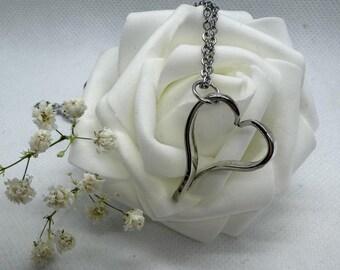 May include: A silver-colored heart pendant necklace. The open heart design hangs from a delicate chain. The necklace is displayed on a large white rose, with small white flowers in the background. Jewelry.