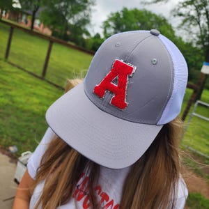 May include: Gray baseball cap with a white mesh back and a red letter "A" on the front. The hat has a curved brim and a snapback closure. The hat is worn by a person wearing a white t-shirt.
