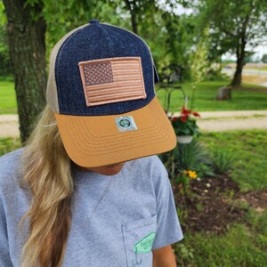 May include: A denim and tan baseball cap with a brown patch featuring an American flag. The cap has a mesh back and a curved brim. The hat is worn by a person wearing a gray t-shirt.