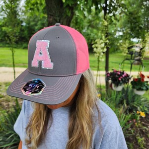 May include: Gray and pink baseball cap with the letter "A" in a sparkly, light pink material. The cap has a mesh back and a curved brim with a pink stitched edge. The brim has a sticker with the text "OC771".