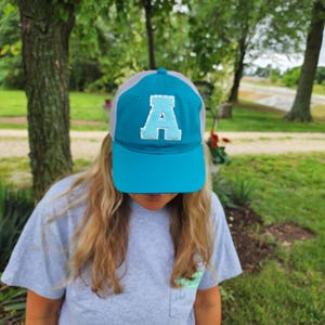 May include: A teal and gray baseball cap with a large, sequined letter "A" on the front. The cap has a curved bill and a mesh back. The person is wearing a gray t-shirt.