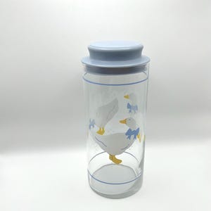 May include: A clear glass jar with a light blue lid and a blue rim. The jar features a design of white geese with yellow beaks and feet, wearing blue bows. The jar is cylindrical in shape.