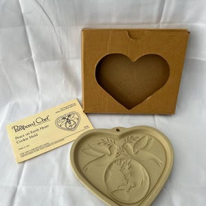May include: A heart-shaped cookie mold with a dove and globe design, along with its original box and instructions. The mold is beige and features the words "Peace on Earth Heart Cookie Mold".