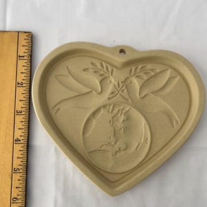 May include: A tan ceramic heart-shaped mould with a design of two doves holding olive branches above a globe. The mould has a small hole at the top for hanging. A wooden ruler is on the left.